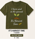 An olive green crewneck t-shirt with a graphic that reads 'I have seed in the ground' and 'It's Harvest Time' around a small sprout illustration. Below the shirt, text reads 'IT'S HARVEST TIME T-SHIRT', 'MILITARY GREEN', and 'S - 5XL'.