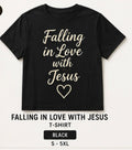 A black short-sleeve T-shirt with the phrase 'Falling in Love with Jesus' and a heart outline printed in beige on the front, shown on a light background with text below reading 'FALLING IN LOVE WITH JESUS T-SHIRT', 'BLACK', and 'S - 5XL'.