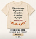 A beige short-sleeve t-shirt with a black typewriter-style quote reading 'Rejoice in hope, be patient in tribulation, be constant in prayer, Romans 12:12' and an orange leaf graphic below. Text underneath the shirt says 'REJOICE IN HOPE ROMANS 12:12 T-SHIRT' and 'NATURAL S - 5XL'.